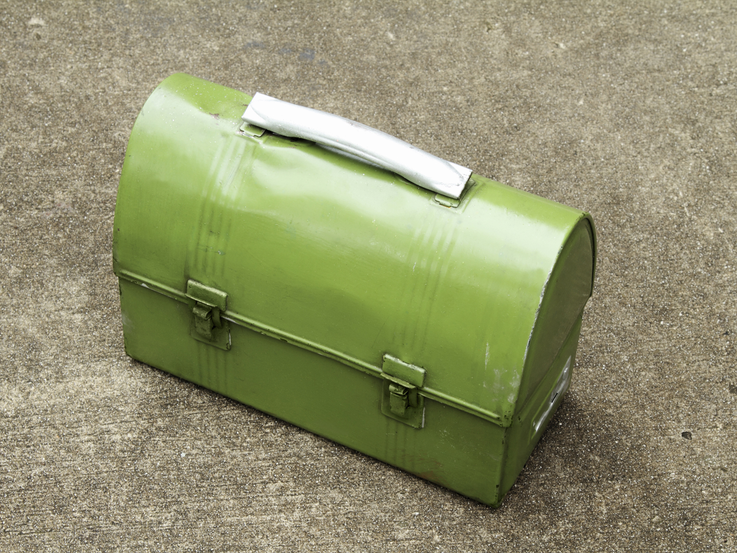 "A green metal lunch box sits on a cement floor. This is an old style work lunch box with some dents, scratches and paint stains."