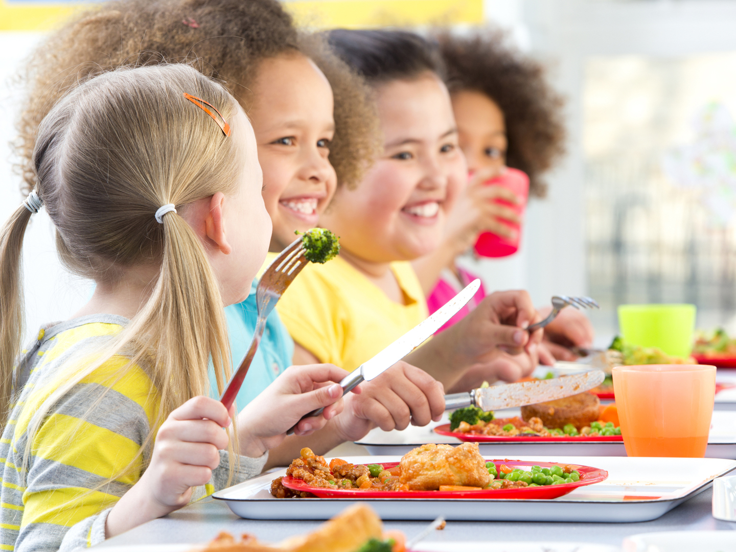 Four children can be seen at a school dinner table with meals infront of them. They are looking at something and smiling, with their cutlery in their hands. One child is having a drink and looking towards his friend, who is looking back at him.