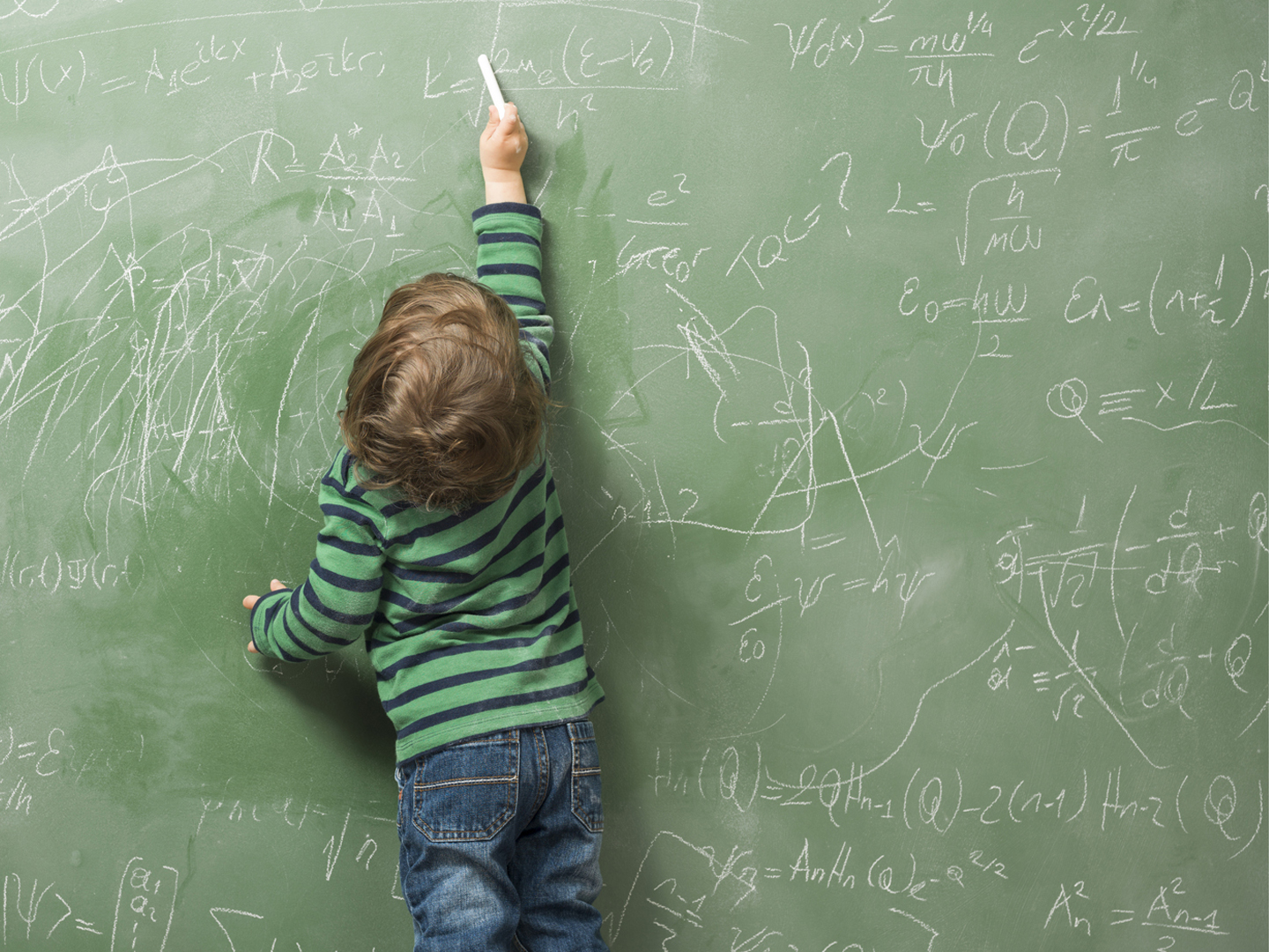 Little boy writing on green blackboard with math formulas written on.He is wearing a rgreen sweater and standing on the left side of frame.The green board is full of mathematics and physics formulas.He is seen in full length and holding chalk in left hand.The photo was shot in studio with a medium format camera Hasselblad H4D.