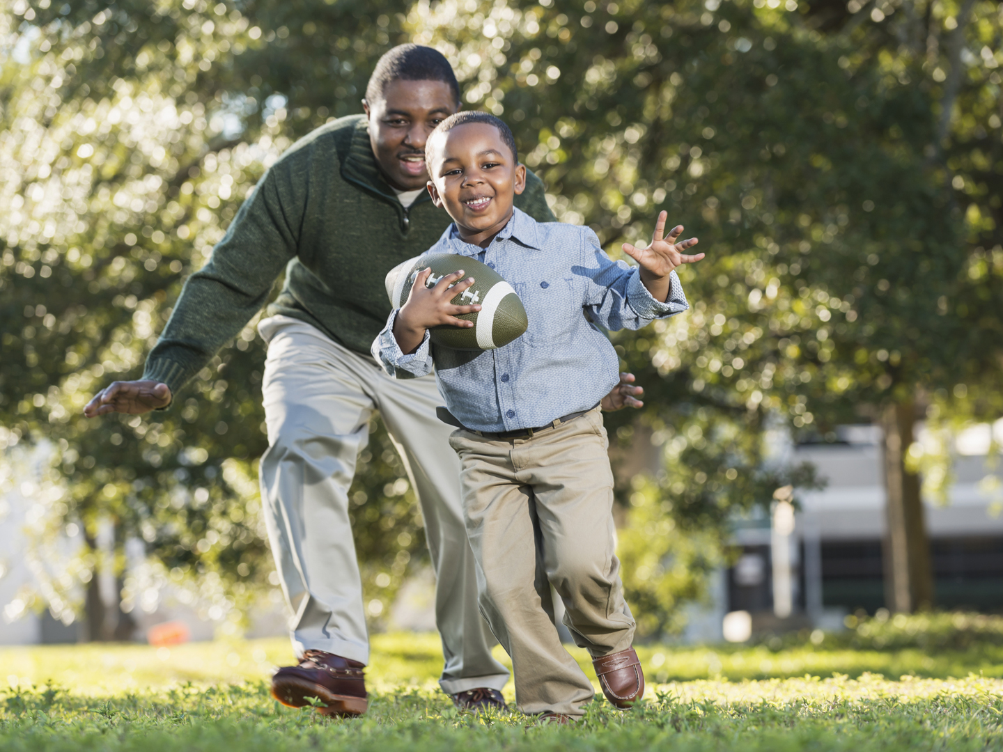 An African American man in his 40s playing football with his 5 year old son. It is a bright, sunny spring day. They are outdoors in the park or the yard, trees out of focus in the background. The boy is holding the ball, running, and his dad is chasing him from behind.