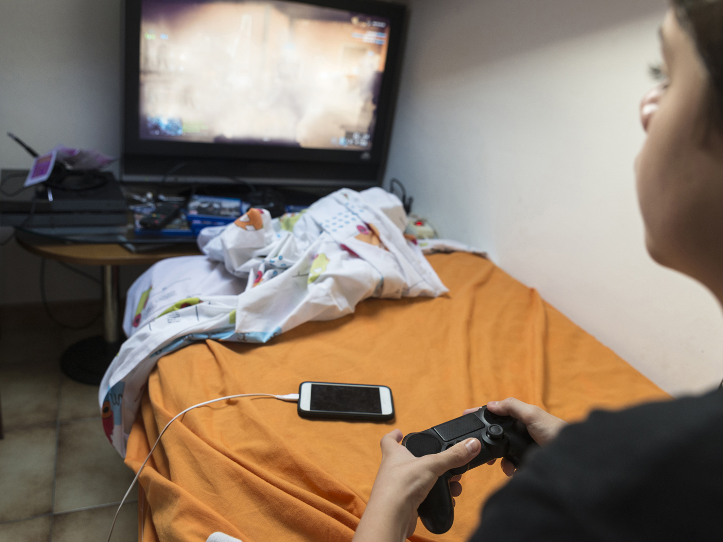 Young boy playing video games sitting on the bed in her room while charging the phone