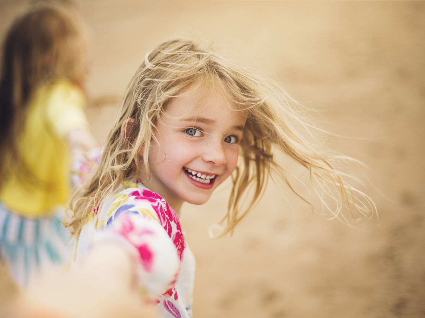 Little girl holding the hand of the photographer that is out of focus in the foreground. From a personal perspective the little girl leads the photographer along the beach and looks back with a big smile. Only the girls face is in focus.