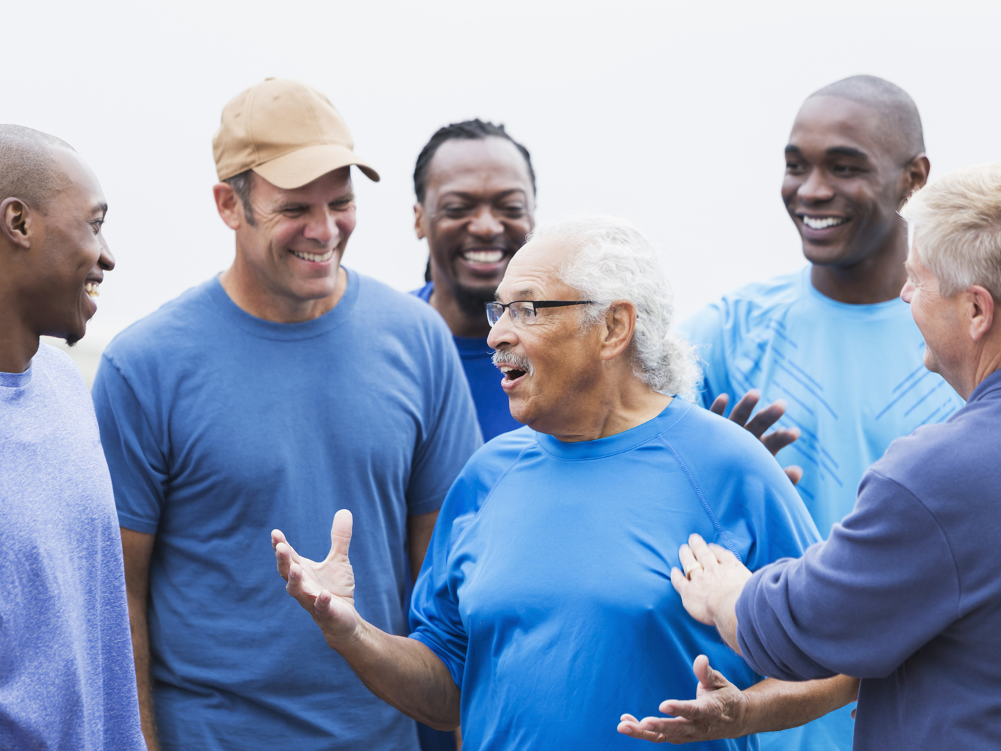 Senior Hispanic man, in his mid 70s, standing with a group of younger male multiracial friends, talking.  He is the center of attention.