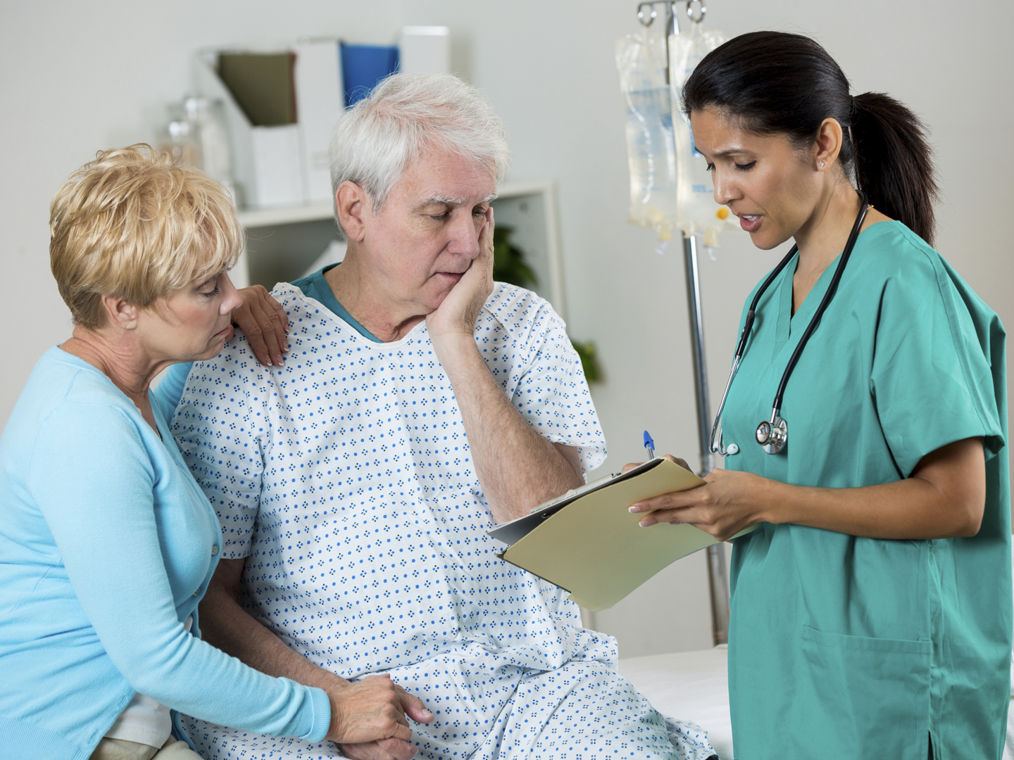 Mature Hispanic doctor or nurse talks with senior patient and his wife about his test results. The man and his wife are sad and he has his hand on his face. They are holding hands. He is connected to iv drip. The nurse or doctor is wearing scrubs and is holding the test results attached to his medcal chart. The man is wearing a hospital gown.