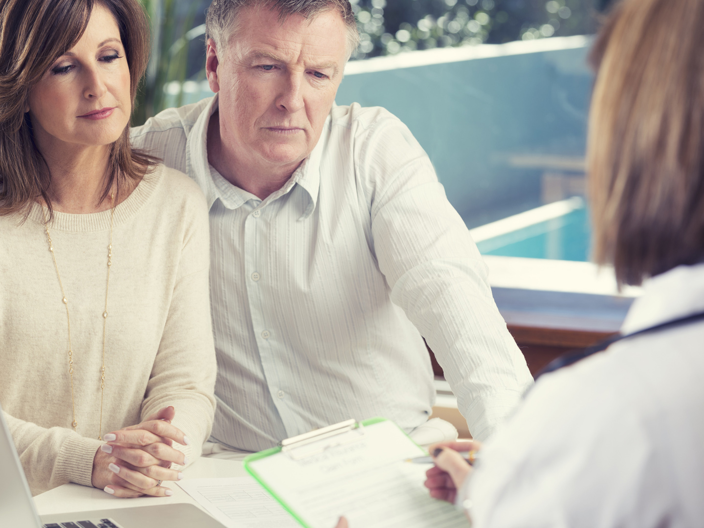 Mature couple with doctor. Both look concerned, worried and upset. Doctor is in foreground holding a clipboard with medical results.