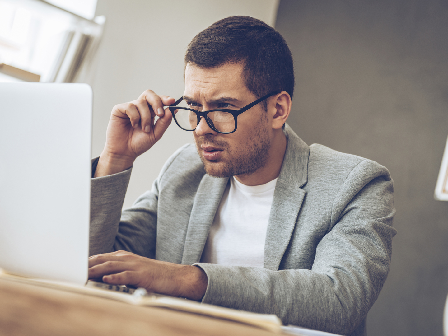 Handsome young man looking at his laptop and adjusting his glasses while sitting at his working place