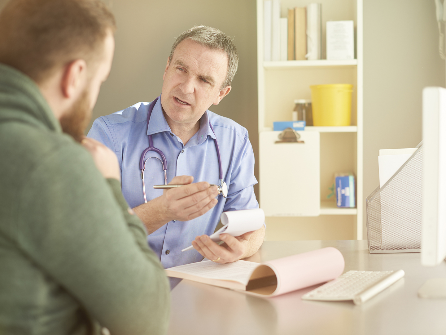 gp explains something to his male patient whilst writing a prescription