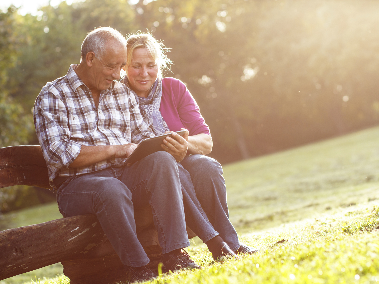 Senior couple sitting on a park bench on sunny autumn day with tablet and relaxing.