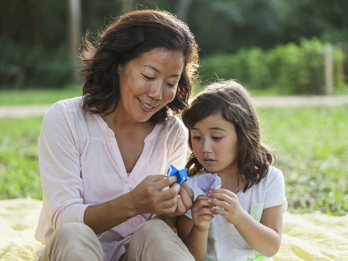 Japanese woman showing her daughter how to fold paper into a flower, doing origami. They are at the park, sitting on a blanket on the grass. The mother has a smile on her face and the little girl is serious, looking down at the paper.