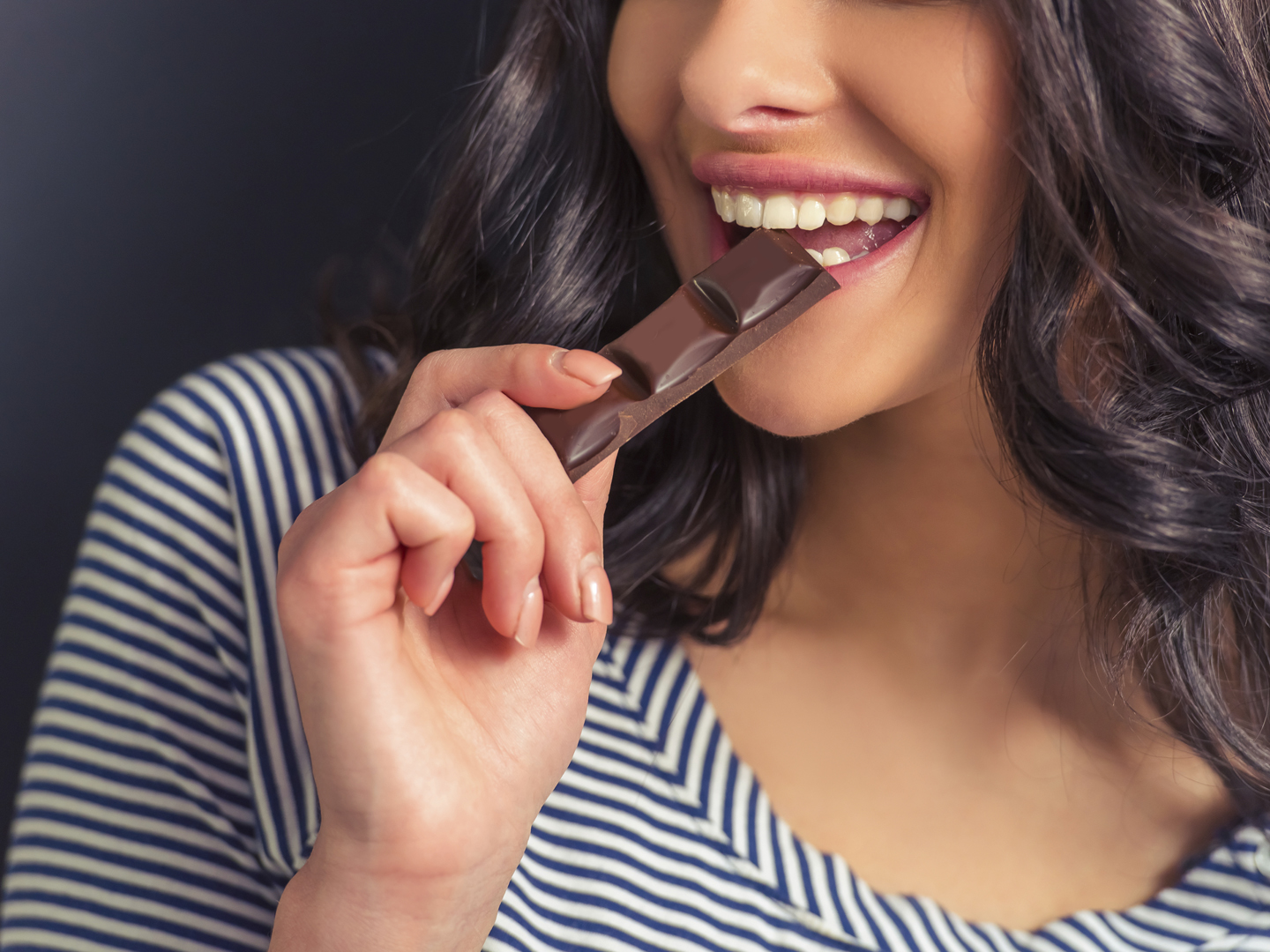 Cropped image of attractive girl eating chocolate and smiling, against dark background