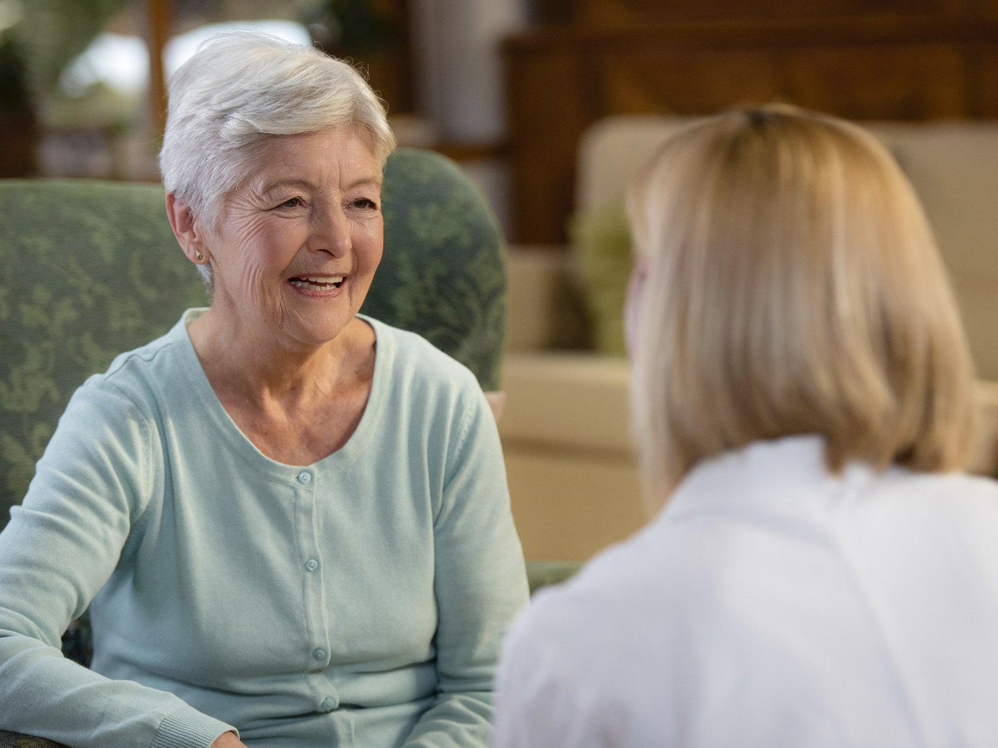 Front view of smiling senior patient sitting in armchair talking with home healthcare nurse or female doctor, rear view.