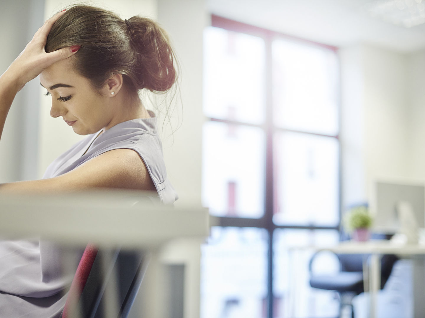 a young office worker looks sad or exhausted at her desk . She hangs her head in her hands . In the background an empty desk is shown defocussed .