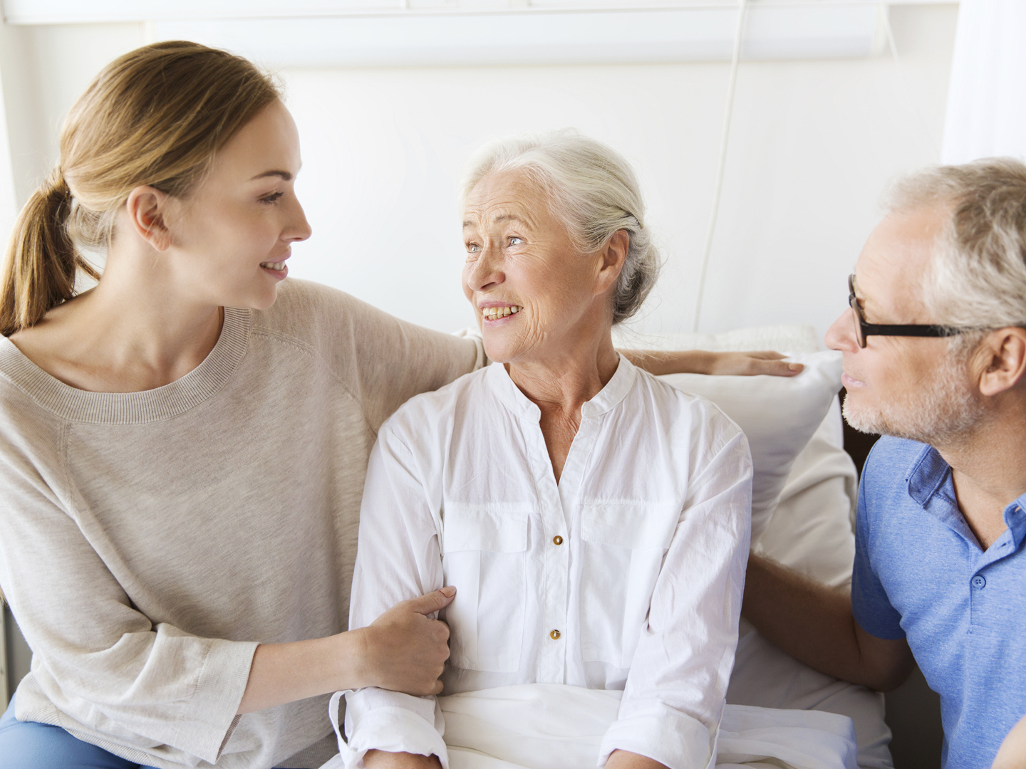 medicine, support, family health care and people concept - happy senior man and young woman visiting and cheering her grandmother lying in bed at hospital ward