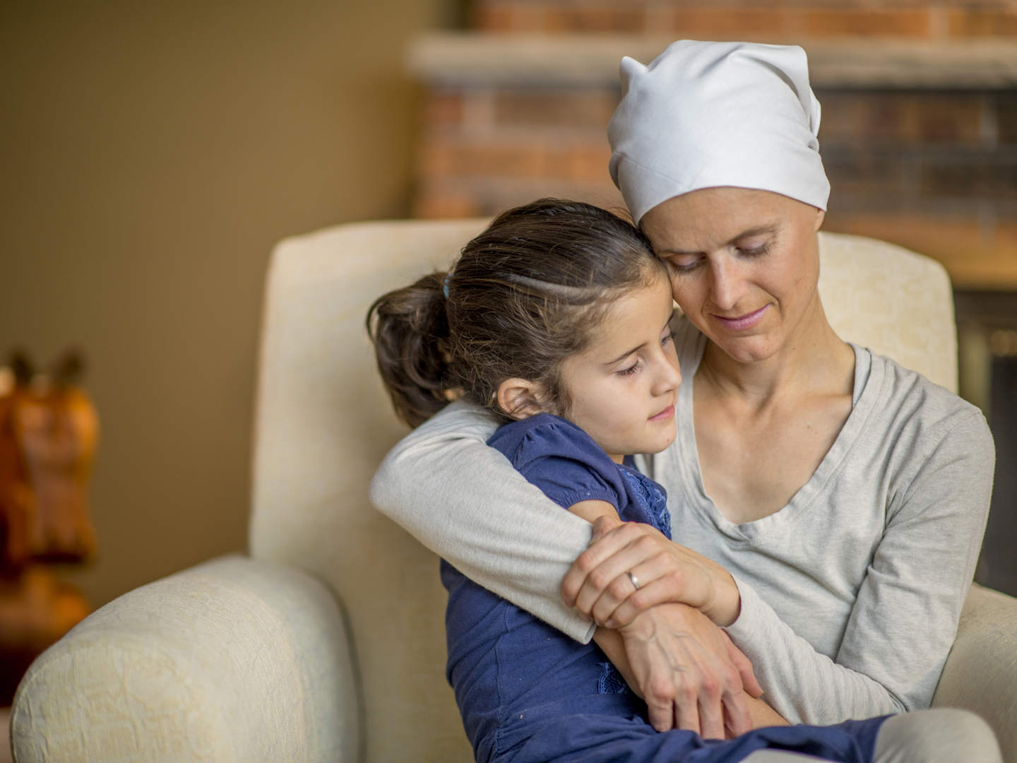 A mother with cancer is sitting in their home and is holding her daughter on her lap.