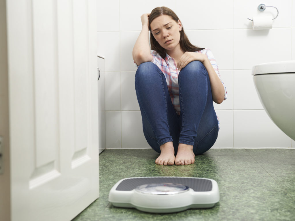 Unhappy Teenage Girl Sitting On Floor Looking At Bathroom Scales