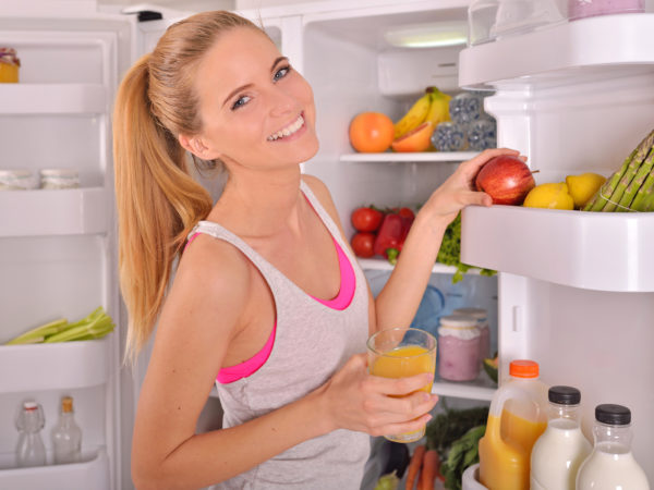 Young beautiful woman standing in the refrigerator door in fitness clothes and drinking orange juice while looking at the camera and smiling.