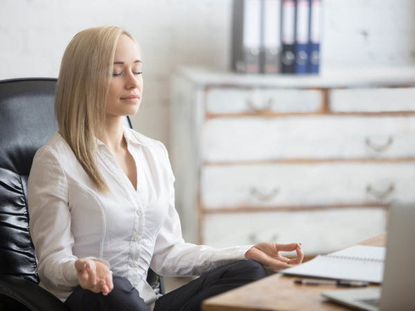 Business and healthy lifestyle concept. Portrait of young office woman sitting cross-legged in easy yoga pose at workplace. Smiling business lady meditating on her break time