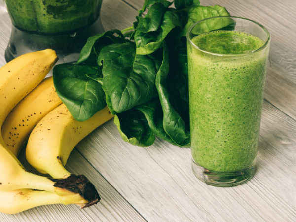 Vitamin Smoothies in a glass on a wooden table. On the table are also banana, spinach and a food processor.