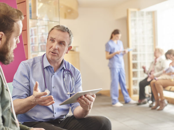 a mature male doctor sits next to a male patient in his waiting room and casually chats through a few things on his digital tablet . In the background a nurse chats to a mother and child and grandmother with her digital tablet .