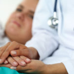 Friendly female doctor hands holding patient hand lying in bed for encouragement, empathy, cheering and support while medical examination. Bad news lessening, compassion, trust and ethics concept