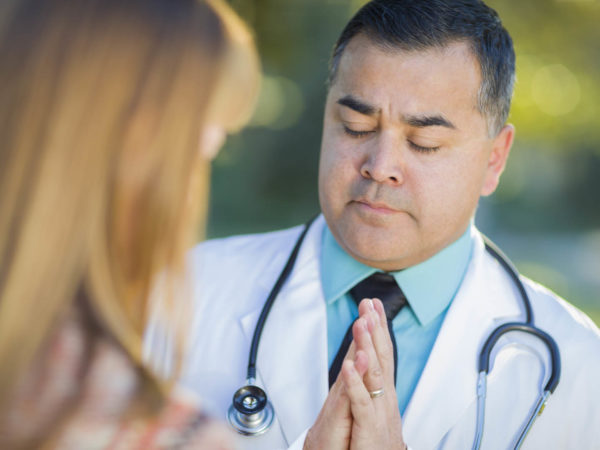 Hispanic Male Doctor Or Nurse Praying With A Patient Outdoors.