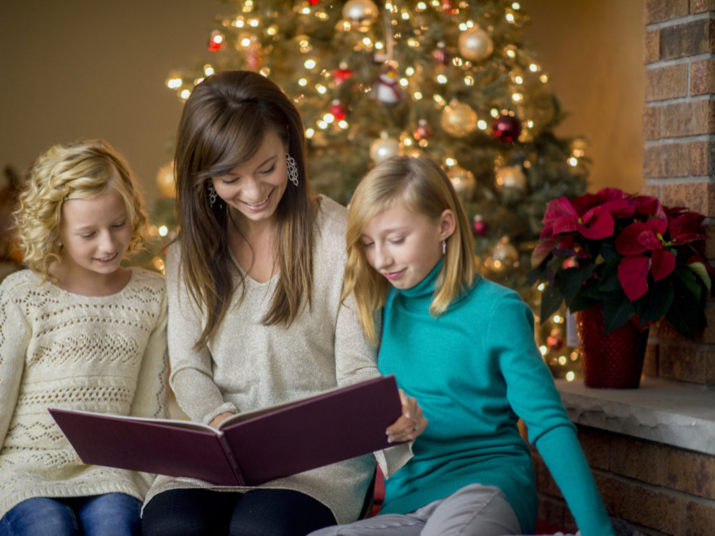 A mother is reading a Christmas story to her two daughters - they are sitting in the living room next to their tree.