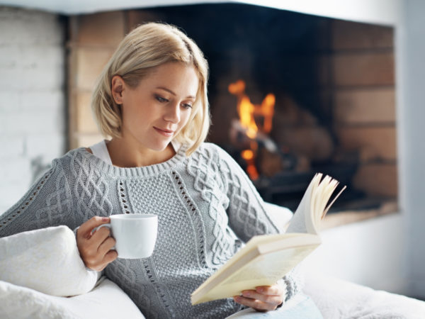 A beautiful young woman reading a book and enjoying a warm beverage near a fireplace