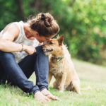 A mature Caucasian woman has relaxing fun playing with her dog in a beautiful park setting.  She lovingly talks to him with a smile on her face.  They rest in the grass, the woman arm over the dog.  Horizontal image with copy space.