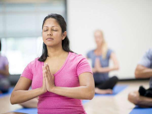 A multi-ethnic group of adults are taking a yoga class together in the gym. They are sitting together and are meditating with their eyes closed on their yoga mats.