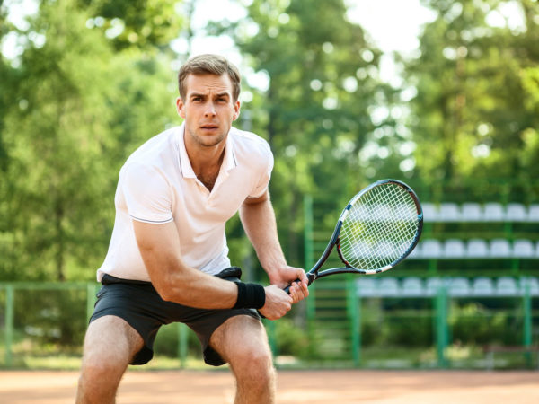 Picture of handsome young man on tennis court. Man playing tennis. Man is ready to hit tennis ball. Beautiful forest area as background