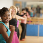 A multi-ethnic group of men and women in a Zumba dance fitness class in an indoor gym studio, standing in a row, wearing exercise clothing, and moving to the music.