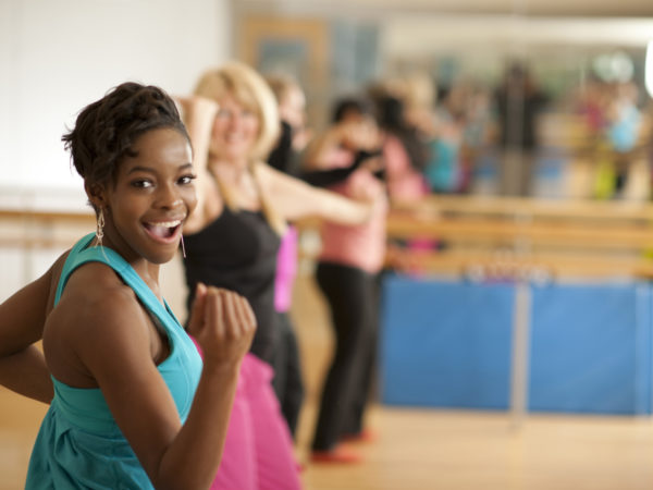 A multi-ethnic group of men and women in a Zumba dance fitness class in an indoor gym studio, standing in a row, wearing exercise clothing, and moving to the music.