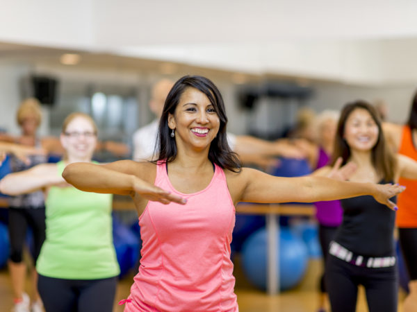 A woman is leading out in a fitness dance class at her indoor studio. She is teaching a multi-ethnic class that is wearing fitness clothing and dancing to the music.
