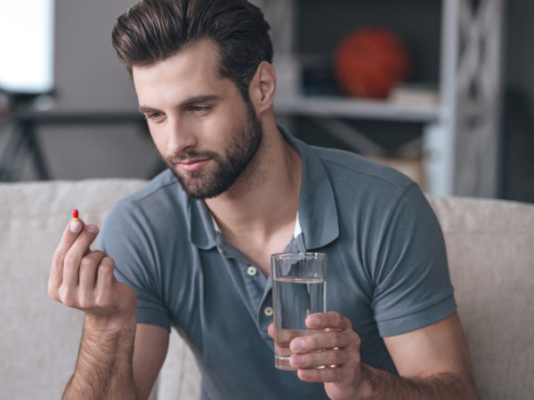 young man holding a glass of water and looking at a pill in his hand while sitting on the couch at home