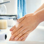 Side view of unrecognizable caucasian woman washing her hands with soap in bathroom. Water is pouring over her hands, visible bluish sink in background.