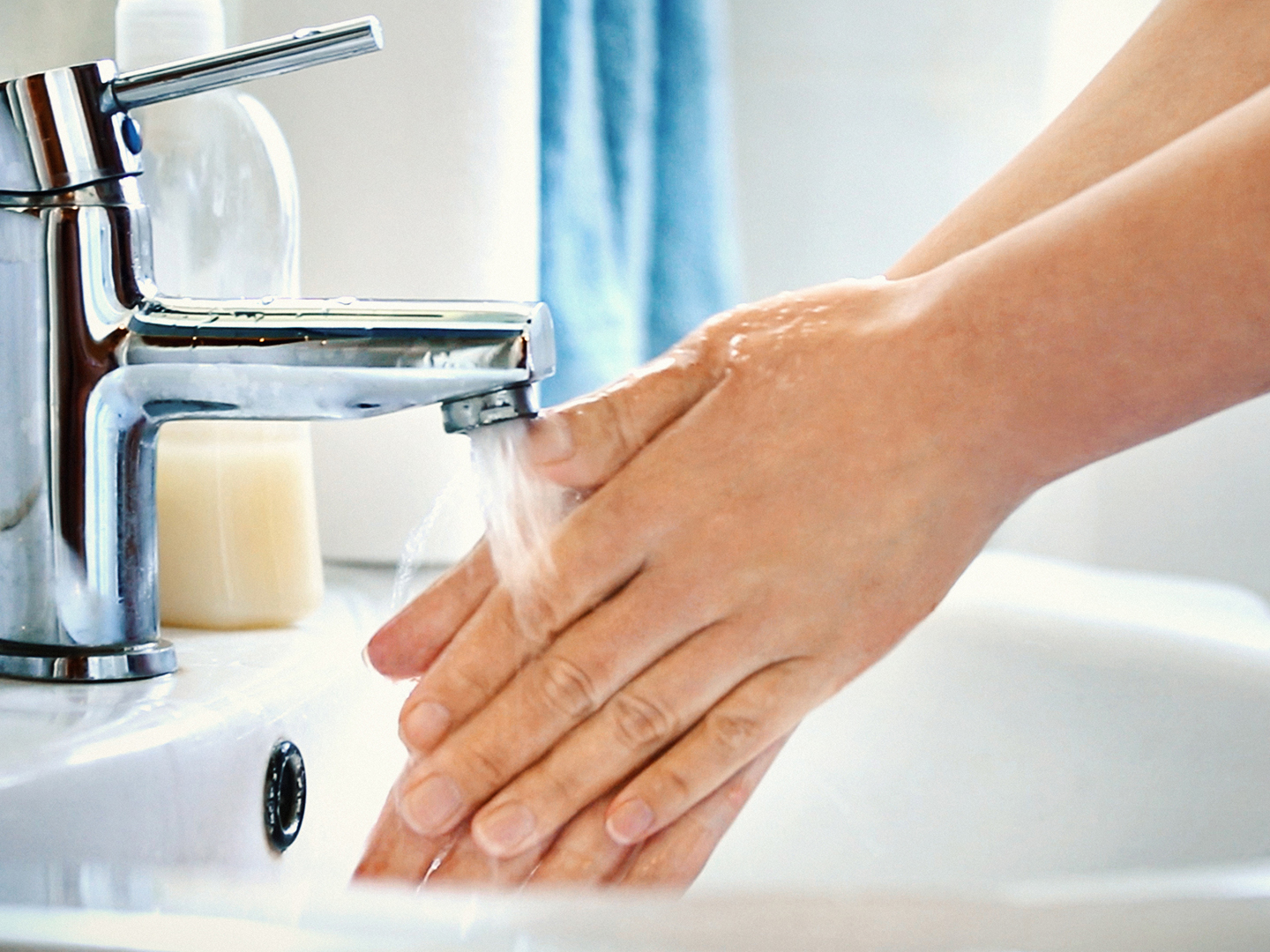 Side view of unrecognizable caucasian woman washing her hands with soap in bathroom. Water is pouring over her hands, visible bluish sink in background.