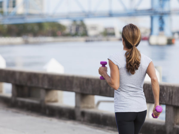 Rear view of mature woman staying fit, running or power walking along the waterfront, carrying dumbbells in her hands.