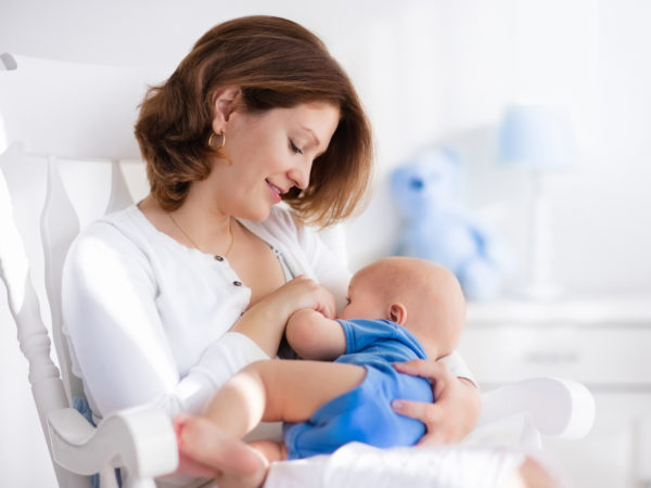 Young mother holding her newborn child. Mom nursing baby. Woman and new born boy in white bedroom with rocking chair and blue crib. Nursery interior. Mother playing with laughing kid. Family at home