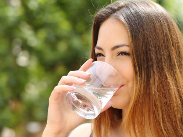 Happy healthy woman drinking fresh water from a glass outdoor with a green background