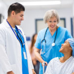 Young adult African American female patient is smiling while talking to mid adult Hispanic male doctor and senior adult Caucasian nurse in hallway of hospital. Nurse is pushing patient in wheelchair toward recovery room after outpatient surgical procedure.