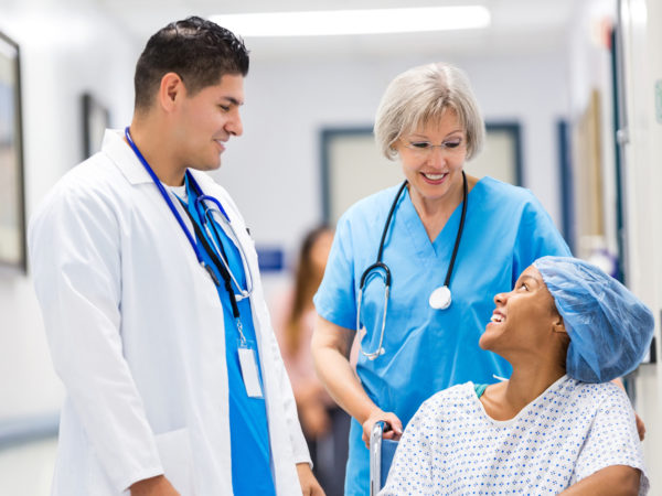 Young adult African American female patient is smiling while talking to mid adult Hispanic male doctor and senior adult Caucasian nurse in hallway of hospital. Nurse is pushing patient in wheelchair toward recovery room after outpatient surgical procedure.