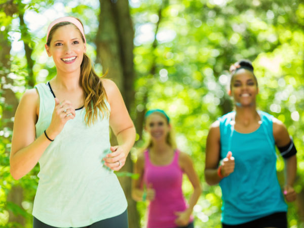 Mid adult Caucasain brunette woman is fitness instructor, leading running club of diverse women. Young adult African American and Caucasian athletic women are following her as they jog or run off road in wooded park on sunny summer day. Women are wearing athletic tank tops and clothing.
