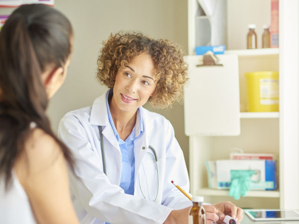 A female doctor sits at her desk and listens to a female patient .  She is wearing blue scrubs and stethoscope and beams a big happy smile as she listens to the young female patient in front of her .