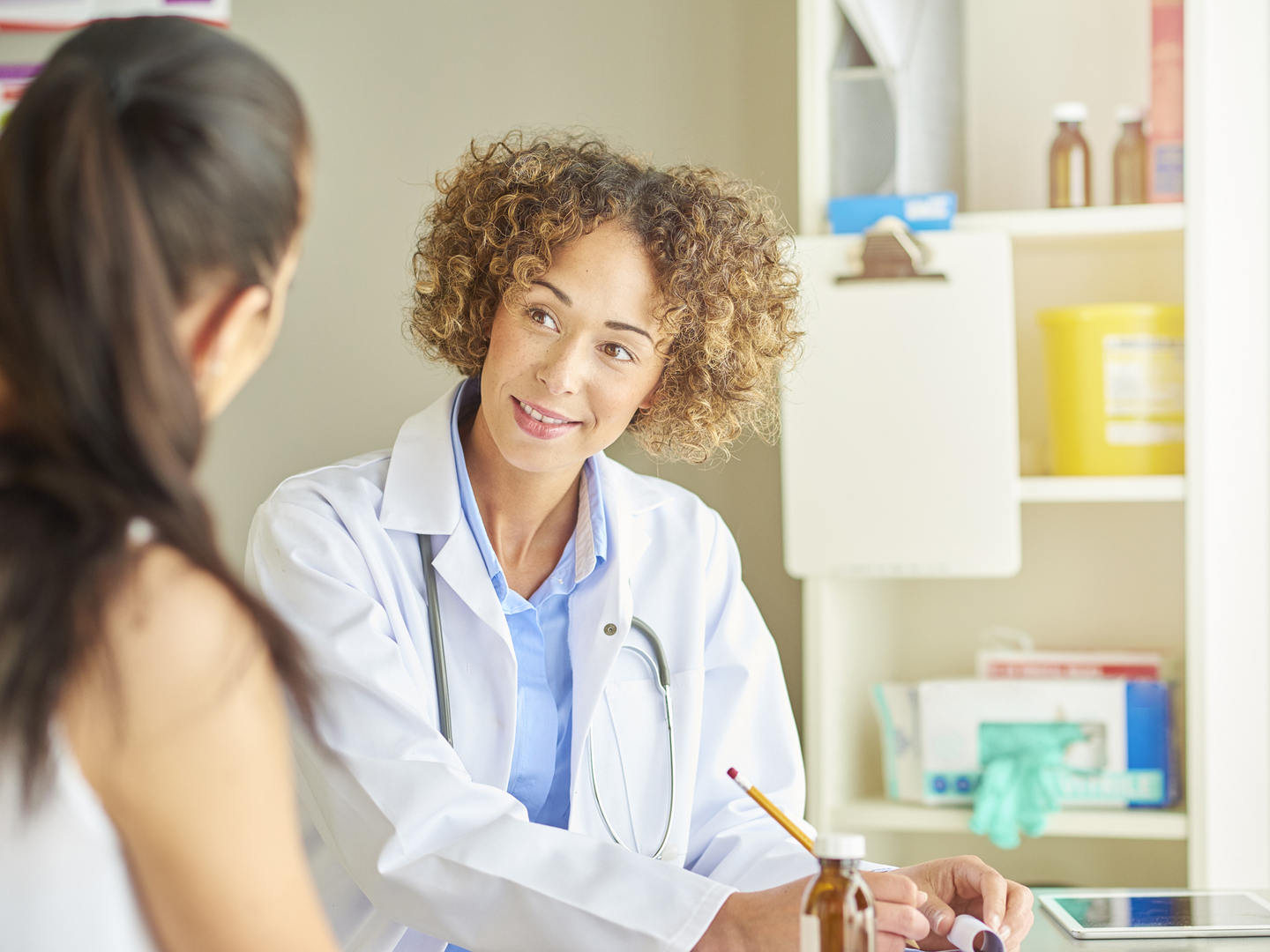 A female doctor sits at her desk and listens to a female patient . She is wearing blue scrubs and stethoscope and beams a big happy smile as she listens to the young female patient in front of her .