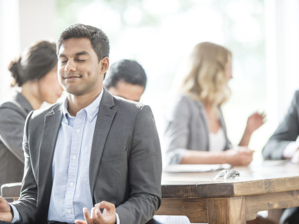 A multi-ethnic group of business professionals are in a a meeting together in the boardroom. One man is meditating peacefully and creating a work life balance.