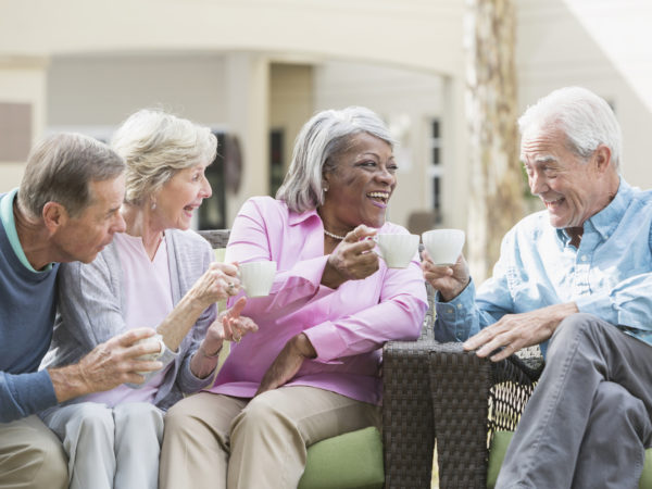 A group of four multi-ethnic seniors sitting together on patio furniture outdoors, talking and smiling, drinking tea or coffee. One of the men and the African American senior woman are toasting with their cups. They are in retirement, relaxed and enjoying spending time with friends.
