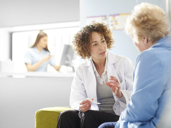 A female pharmacist sits with a senior female patient in the pharmacist consultation area and discusses her prescription and choice of medication. In the background a father and daughter stand at the dispensing counter and are served by a female pharmacy assistant .