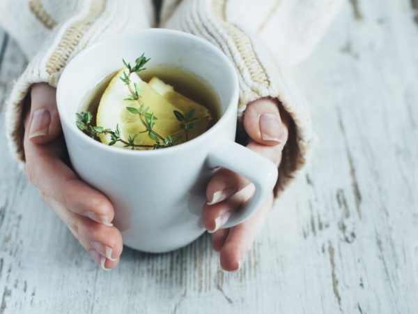 Human hands holding cup of tea with thyme herb and lemon slices on a wooden table,.