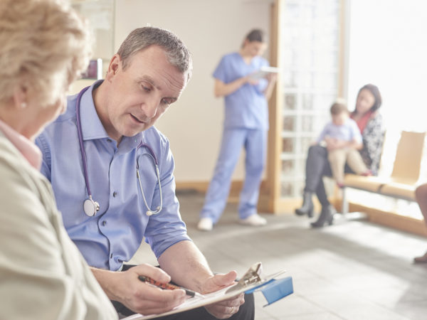 a mature male doctor sits next to a senior patient in his waiting room and casually chats through his factsheet . In the background a nurse chats to a mother and child with her digital tablet .