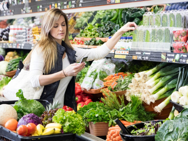 Woman with cart shopping in grocery store in the produce section. Using smart phone